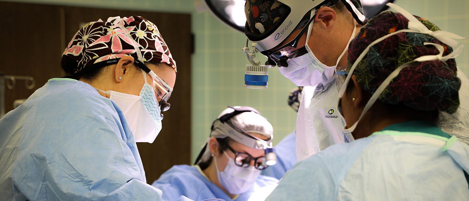 Three surgeons in masks, magnifying loupes and headlights lean in over the operating field during a microvascular reconstruction.