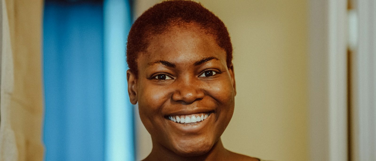 A woman with short, cropped auburn hair smiles warmly in a softly lit room.
